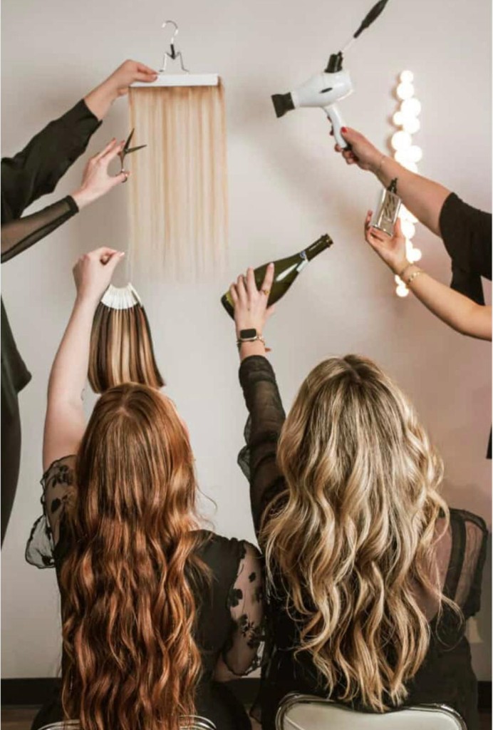 Editorial salon scene: two models with long wavy auburn and blonde hair, styling tools, extension weft, and color swatches